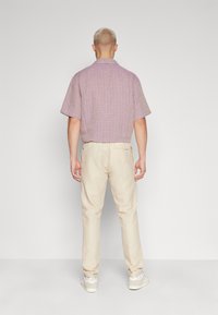 Korte mouwen, geknoopte blouse in licht paars met een gestructureerd patroon, gecombineerd met beige broeken en witte sneakers. Effen achtergrond.