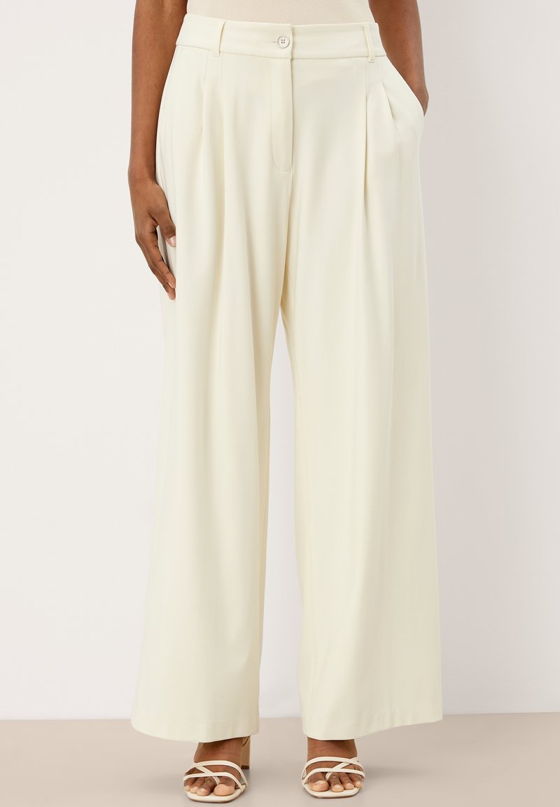Pantalon crème taille haute, jambes larges, avec plis devant, poches latérales, fermeture éclair et bouton, porté avec des talons blancs à brides.
