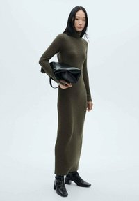 Robe tricotée moulante vert olive avec un haut col roulé, des manches longues et une silhouette droite. Mannequin tenant un sac noir avec une bandoulière.