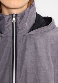 Lehká šedá bunda s vysokým límcem, černou síťovinou uvnitř a předním zipem. Obsahuje strukturovanou látku pro prodyšnost.