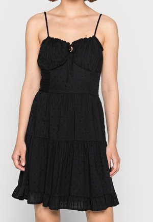 Femme portant une robe noire sans manches avec des bretelles fines, un buste froncé avec un lien, et un ourlet à volants au-dessus du genou.