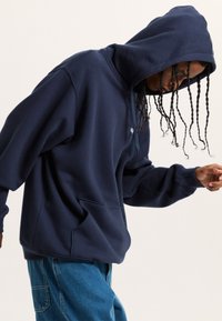 Felpa blu navy con tasca frontale, cappuccio con cordini e polsini elasticizzati. Indossata con jeans blu, presenta un fit casual e rilassato.