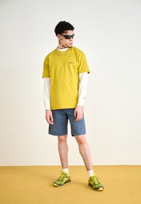 Gelbes Baumwoll-T-Shirt, das über einem weißen Langarmshirt getragen wird, mit blauen Denim-Shorts und grün-schwarzen Sportschuhen mit reflektierenden Akzenten.