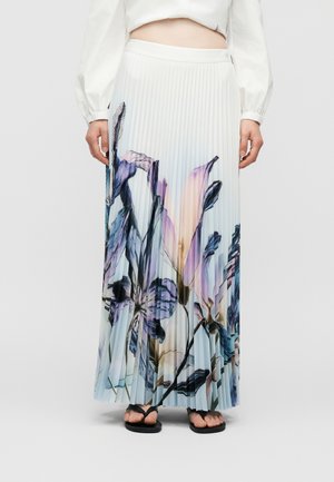 Γυναίκα που φοράει λευκή πλισέ μακριά φούστα με μεγάλα μοβ και μπλε λουλουδάτα σχέδια, σε συνδυασμό με μαύρα σανδάλια και λευκό κοντό πουκάμισο.