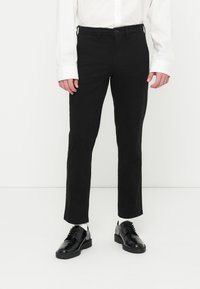 Pantalon noir à jambes droites en tissu lisse, avec fermeture à boutons sur le devant et poches latérales. Associé à des chaussures noires brillantes.