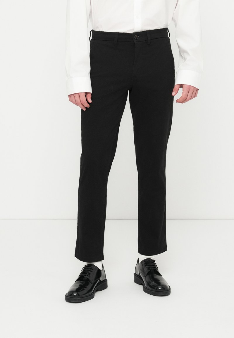 Pantalon noir à jambes droites en tissu lisse, avec fermeture à boutons sur le devant et poches latérales. Associé à des chaussures noires brillantes.