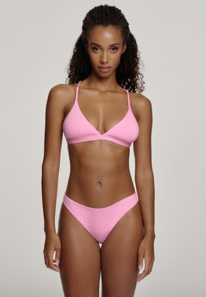 Kvinde med krøllet hår iført en pink bikini med to dele stående mod en ensfarvet lys baggrund, vendt fremad.