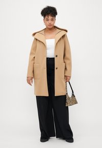 Beige ullfrakk med stor hette, framfickor og svarte knapper, kombinert med svarte vide bukser og en liten veske med zebra-mønster.