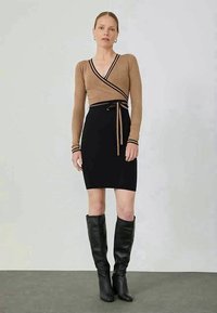 Haut cache-cœur en maille marron clair avec bordure noire, associé à une jupe noire ajustée. Des bottes en cuir noires hautes jusqu'au genou complètent la tenue.