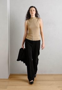 Bežinė, megzta be rankovių palaidinė su aukštu kaklu; derinama su juodomis plačiomis kelnėmis. Modelis laiko juodą švarką, minimalistinis stilius, lygus paviršius.