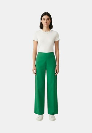 Donna in piedi di fronte, indossa una camicia bianca a maniche corte, pantaloni verdi a gamba larga, scarpe da ginnastica bianche e una sottile collana d'oro.