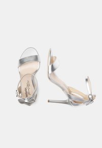 Sandales à talons hauts argentées avec une finition métallique brillante, bride de cheville ajustable, et un petit nœud décoratif au talon. Conception à bout ouvert.