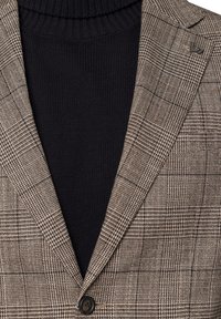 Brązowa marynarka w kratę z czarnym golfem pod spód; marynarka ma teksturowany materiał, gładki guzik i wyraźne klapy.