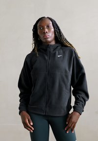 Czarna bluza z kapturem Nike z zamkiem, wykonana z materiału polarowego, z wysokim kołnierzem i haftowanym logo na lewej piersi. Gładka tekstura, luźny krój.
