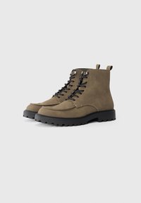 Botas de tobillo de ante verde oliva con cordones negros, suela de goma y lengüeta de tela en el talón, que presentan una punta redondeada y una textura resistente.