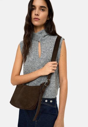 Kobieta ubrana w bezrękawnik z szarego dzianego materiału i ciemnoniebieskie dżinsy, trzymająca ramię ciemnobrązowej zamszowej torby na ramię.