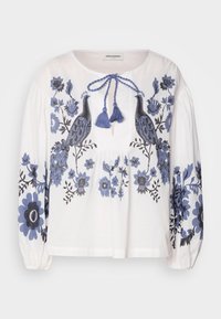 Blouse blanche à manches longues avec broderie florale bleue et motif paon, dotée d'un col rond et d'une fermeture à lacets avec pompons bleus.