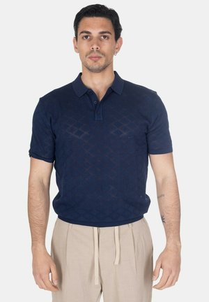 Uomo che indossa una polo blu navy a maniche corte con motivi a diamante e pantaloni beige con coulisse, in piedi su sfondo bianco.