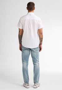 Witte shirt met korte mouwen en een rugpasse, gecombineerd met lichtblauwe jeans en witte sneakers. Heeft een strak, minimalistisch ontwerp en pasvorm.
