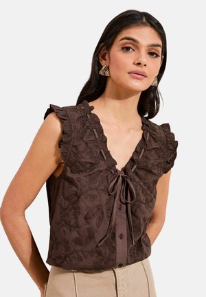 Femme portant un chemisier sans manches marron foncé brodé avec des épaules volantées et un lien au niveau du col, associé à un pantalon beige clair.