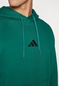Hoodie vert en tissu doux avec une poche avant, arborant un logo Adidas noir et des cordons assortis. Design simple et coupe décontractée.