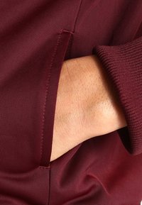 Bordová tkanina s hladkým povrchem má kapsu, do které je zasunuta ruka, zvýrazňující prošívání a elastické manžety.