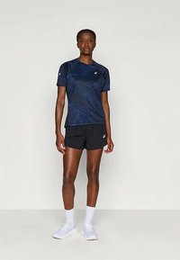 Nais inimene seisab seljas tumesinine mustriga lühikeste varrukatega särk, mustad šortsid, valged sokid ja valged sportlikud jalatsid, ühtlase tausta ees.