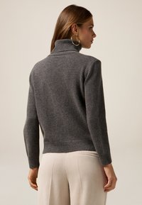 Suéter gris de cuello alto con puños y dobladillo acanalados, que presenta una tela suave y texturizada. Modelo vistiendo pantalones plisados de color beige claro.