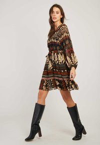 Femme marchant dans une robe à motifs arrivant au genou et des bottes noires à talons hauts contre un fond uni.