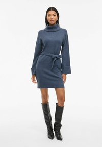 Abito in maglia blu scuro con colletto alto, maniche lunghe e una cintura coordinata in vita. Indossato con stivali neri al ginocchio.