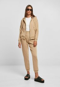 Beige hettegenser med kengurulommer og snor, sammen med matchende joggebukser. Modellen har på seg svarte sandaler med tykke såler.