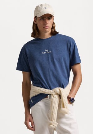 Marineblauwe katoenen t-shirt met witte geborduurde tekst "Polo Ralph Lauren," gecombineerd met een crème kabelgebreide trui die om de taille is geknoopt.
