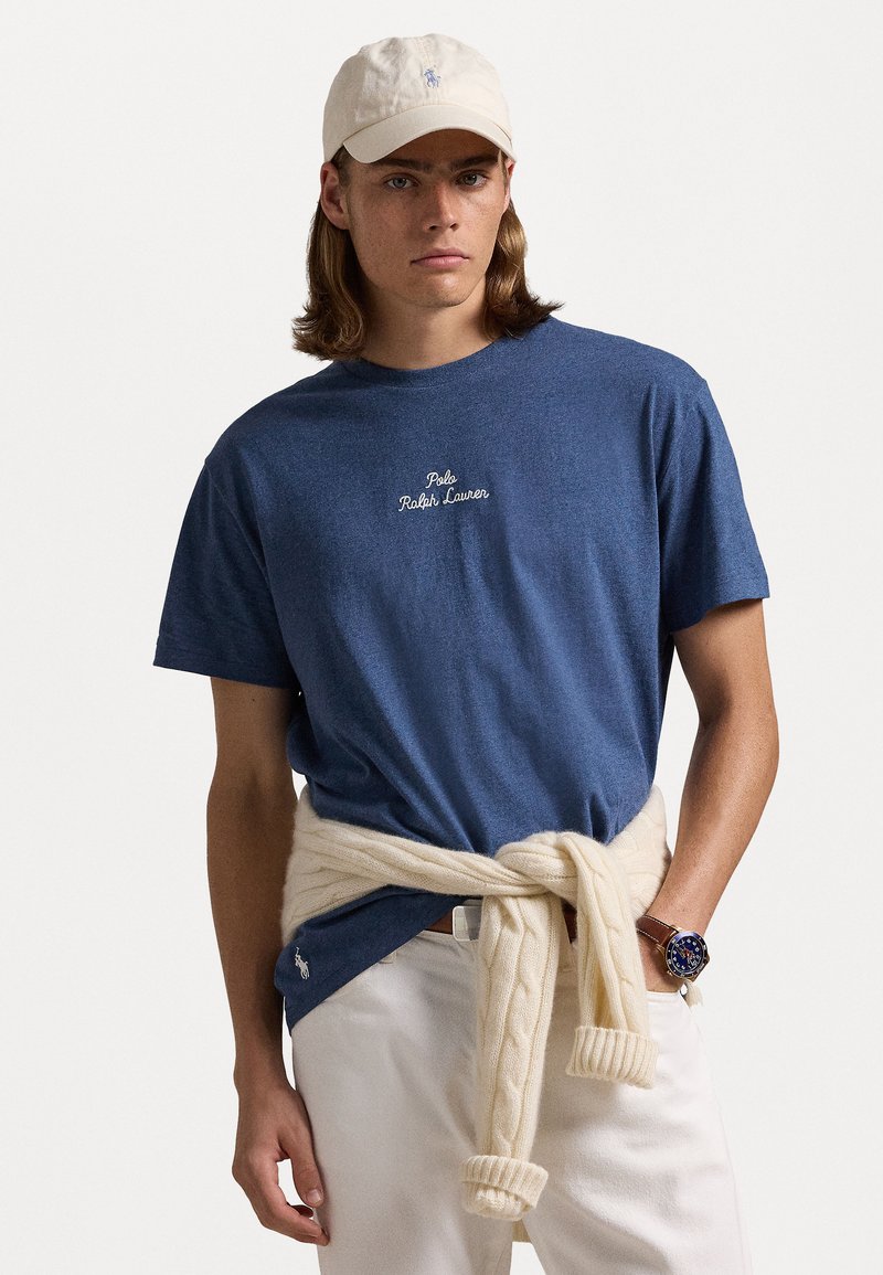 Marinblå bomulls t-shirt med vit broderad text "Polo Ralph Lauren," i kombination med en krämfärgad kabelstickad tröja knuten runt midjan.