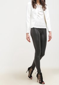 Bílý texturovaný blazer, top s šněrováním v světle barvě, černé přiléhavé legíny a černé sandály na vysokém podpatku se zlatými detaily.