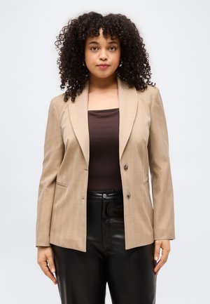 Kobieta o kręconych włosach ubrana w beżową marynarkę na ciemnobrązowej bluzce i czarnych skórzanych spodniach, stoi na jednolitym jasnym tle.