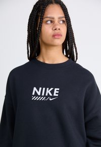 Zwarte sweatshirt met ronde hals, voorzien van het witte "NIKE" logo en streepdesign op de voorkant. De stof lijkt zacht met een ontspannen pasvorm.