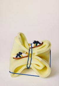 Dos zapatillas blancas con rayas verdes, amarillas y azul marino colocadas sobre espuma amarilla retorcida, sujetas con una cuerda azul sobre un fondo liso.