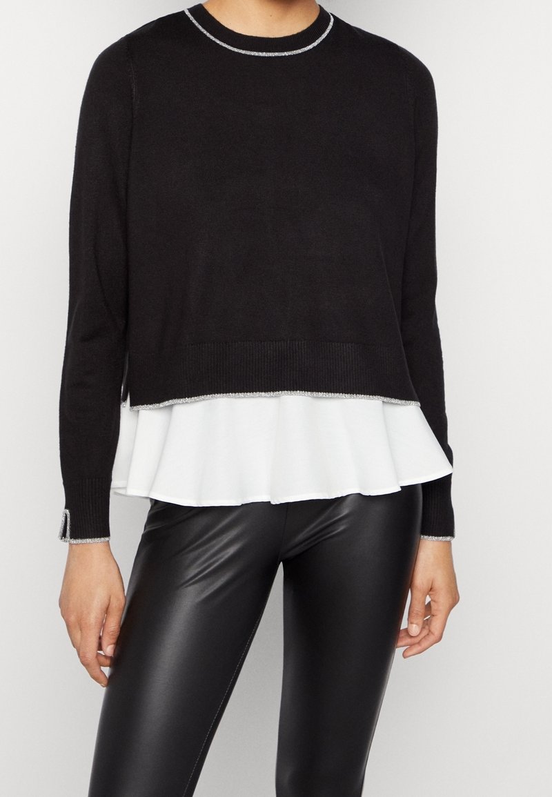 Femme portant un pull noir superposé avec un ourlet volanté blanc, associé à des leggings en simili cuir noir, coupés à hauteur de mi-cuisse.