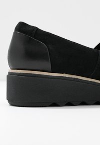 Chaussure à talon compensé noire avec tige en daim, contrefort de talon lisse et bordure beige entre la semelle et la tige.