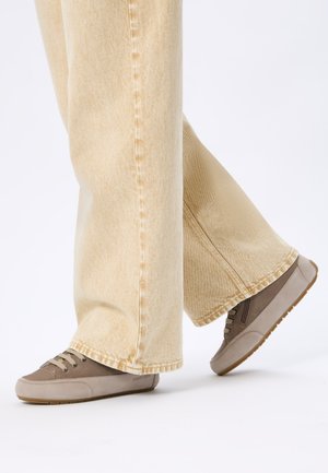 Legs wearing beige wide-leg jeans and light brown casual sneakers stepping on a white floor.