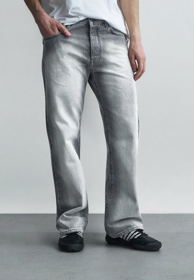 Homme portant un jean gris délavé à jambes droites, un T-shirt blanc et des baskets noires, debout sur un sol gris contre un mur clair.