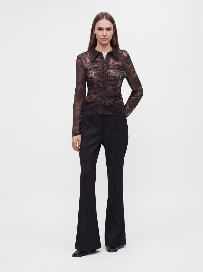 Une femme porte un chemisier noir en dentelle transparente, boutonné sur le devant, et un pantalon noir évasé, sur un fond de studio blanc uni.