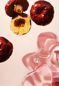 Tørkede røde frukthalvdelar, som viser innvendig tekstur, sammen med hele frukter og klare væskedråper på en lys rosa overflate.