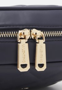 Námořnická prošívaná taška s dvojitým zlatým zipem a vyraženými štítky s názvem značky. Hladký materiál s elegantním, strukturovaným designem.