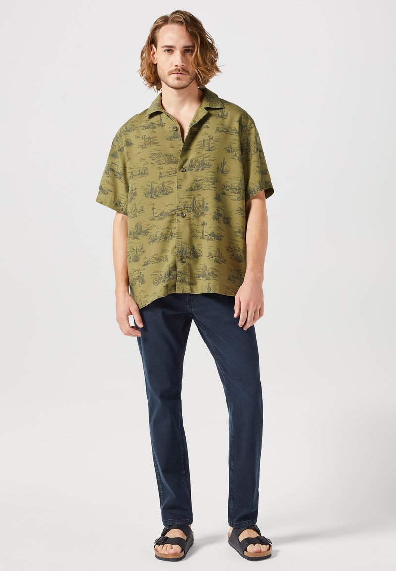 Camisa de manga corta verde oliva con estampado de paisaje negro, combinada con vaqueros azul oscuro y sandalias negras. Cierre de botón simple, corte relajado.