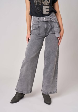 Femme portant un jean gris taille haute à jambes larges avec poches plaquées devant, une ceinture en tissu assortie, des bottes noires et un t-shirt noir.
