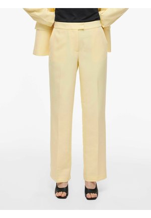 Kvinde iført cremefarvede bukser med højt liv og lige ben, kombineret med sorte hælede sandaler med åben tå, stående med hænderne i lommerne.