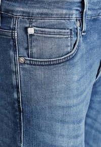 Kék farmernadrág klasszikus öt zsebes dizájnal, enyhén kopott textúrával és fém gombbal a deréknál.