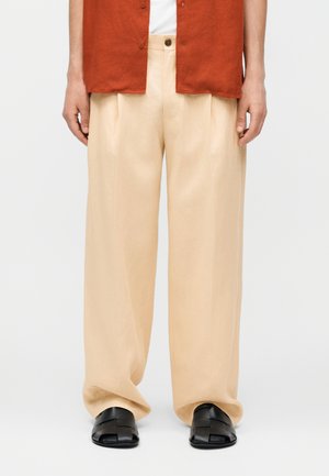 Person iført løse beige bukser, sorte flettede sandaler, hvid skjorte og rustfarvet knaplukket kortærmet top, stående mod en ensfarvet baggrund.