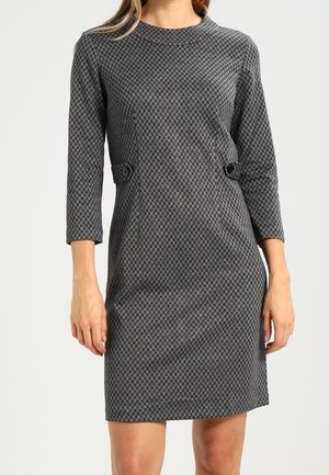 Kobieta w dopasowanej, szarej sukience o długości do kolan, z długim rękawem, ozdobionej drobnym wzorem w romby i guzikami na wysokości talii.